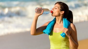 Fitness beautiful woman drinking water and sweating after exercising on summer hot day in beach. Female athlete after work out.