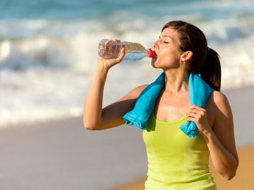 Fitness beautiful woman drinking water and sweating after exercising on summer hot day in beach. Female athlete after work out.